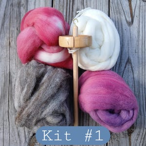 May include: A spinning kit with four bundles of colorful wool roving in pink, white, gray, and purple, along with a wooden spindle. The text "Kit #1" is displayed in a blue box. The items are arranged on a weathered wooden surface.