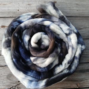 May include: A spiral of dyed wool roving in shades of white, blue, and brown. The fiber is arranged in a circular pattern, creating a visually appealing texture. The wool is likely used for spinning or felting projects.