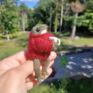 May include: A small, handcrafted knitted frog figurine with a textured green body and black eyes, wearing a fuzzy, deep red long-sleeved sweater. The frog is holding a tiny white ceramic mug in its hands, with its legs dangling downward.