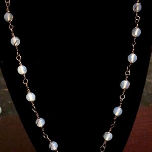 Op de afbeelding: Een delicate ketting gemaakt met witte opaal kralen en koperdraad. De ketting wordt getoond op een zwarte fluwelen mannequinbuste.
