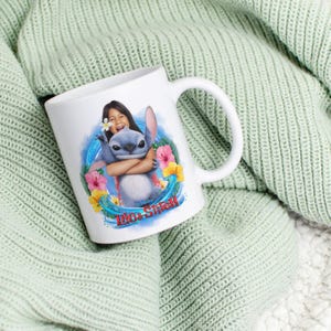 May include: White ceramic mug featuring a colorful graphic of Lilo & Stitch. The design includes Lilo hugging Stitch, surrounded by tropical flowers and the words "Lilo & Stitch" in a stylized font. The mug has a curved handle.