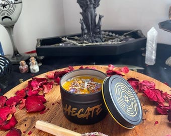May include: A black candle jar with a gold label that reads "HEcate" and a gold lid with a star and circle design. The candle is surrounded by red rose petals and a wooden stick with dried herbs and rose petals.