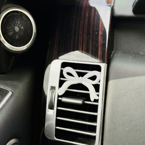 May include: A white bow-shaped car air vent accessory. The bow is positioned on a silver air vent, set against a dark interior with wood grain accents. The image is a close-up, highlighting the accessory's design.