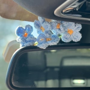 May include: A cluster of crocheted blue flowers with yellow centers, resting above a car's rearview mirror. The handmade flowers add a touch of color and charm to the vehicle's interior. The flowers are a light blue color.