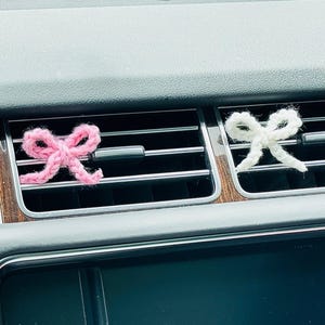 Pode incluir: Dois ambientadores de carro em forma de laço, feitos à mão em malha. Um é rosa, o outro é branco. Estão colocados nas saídas de ar do painel cinzento de um carro, com incrustações de madeira.