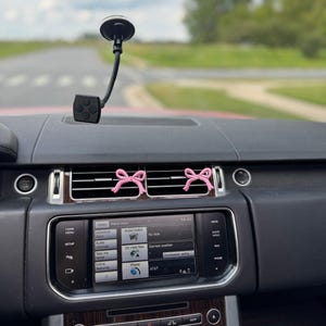 May include: Interior car dashboard with a black phone mount and two pink bow-shaped air vent decorations. The dashboard has a digital display with menu options and a wooden panel.