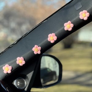 May include: Five pink crocheted flowers with yellow centres are attached to the black interior of a car. The flowers are arranged in a row along the top of the car's interior.