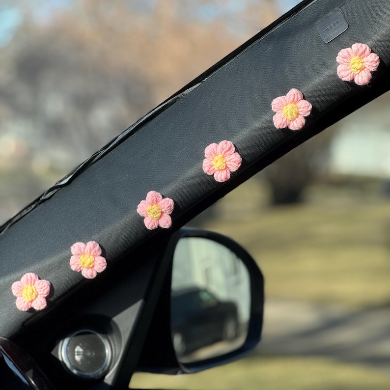May include: Five pink crocheted flowers with yellow centres are attached to the black interior of a car. The flowers are arranged in a row along the top of the car's interior.