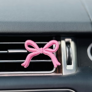 May include: A pink embroidered bow car air vent clip. The bow is centered on the black dashboard, above the air vent. The time 14:45 is displayed on the dashboard. The car interior is black.