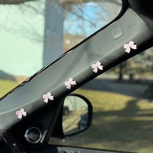 May include: Five small pink bows are attached to the inside of a car's sun visor. The bows are made of a delicate, crocheted material.