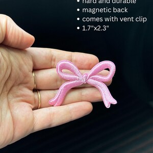 May include: A pink embroidered bow patch, approximately 1.7" x 2.3", with a magnetic back and vent clip. The patch is hard and durable. The image shows the bow held in a hand.