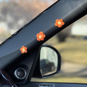 May include: Three crocheted flowers in orange with yellow centres are attached to the sun visor of a car. The flowers are arranged in a row, with the largest flower in the middle.