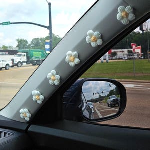 May include: A car's interior with a row of crocheted white daisy flowers with yellow centres attached to the window frame. The flowers are arranged in a line, starting from the top of the frame and going down.