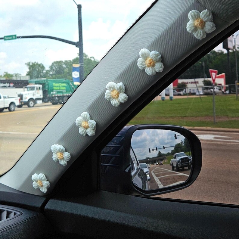 May include: A car's interior with a row of crocheted white daisy flowers with yellow centres attached to the window frame. The flowers are arranged in a line, starting from the top of the frame and going down.