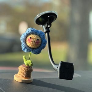 May include: A blue crocheted flower with a smiling face in a brown pot attached to a black car phone holder.