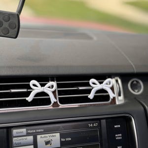 May include: Two white bow-shaped car air fresheners clipped to the air vents of a car's dashboard. The dashboard is black, and the car's infotainment screen displays various options. The time is 14:47.