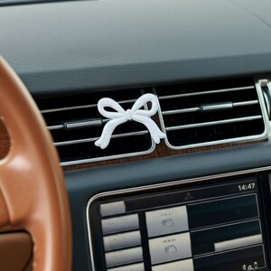 May include: A white bow-shaped car air freshener clipped to a car vent. The bow is centered on the vent, with a brown leather steering wheel and dashboard visible in the background. The car's display screen shows the time.