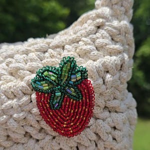 May include: Cream-colored crocheted handbag with a beaded strawberry applique. The strawberry is red with green leaves and a yellow beaded border. The bag has a braided handle and a textured surface.