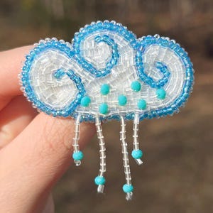 May include: A handmade cloud-shaped brooch. The brooch is made of white and blue beads, with turquoise beads representing raindrops. The cloud has a detailed, textured appearance, with a light blue beaded outline. The brooch is a unique accessory.