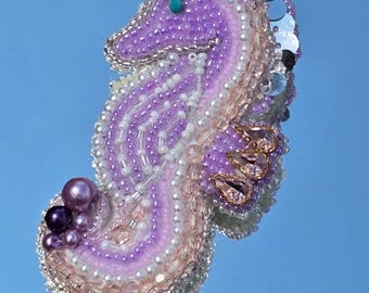 Puede incluir: Broche de caballito de mar hecho a mano, adornado con cuentas en tonos lavanda, blanco y rosa. El diseño incorpora lentejuelas y pequeñas piedras preciosas. El broche tiene un borde plateado y una pequeña cuenta turquesa para el ojo.