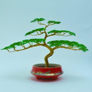 Puede incluir: Escultura de árbol bonsái hecha a mano con alambre verde y dorado, colocada en una maceta de cerámica roja. El árbol tiene un tronco retorcido y follaje verde en capas, creando una apariencia de árbol en miniatura.