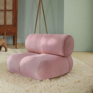 May include: A pink, low-profile armchair with a rounded back and seat. The chair is made of a textured fabric and sits on a cream-colored, shaggy rug. The design is modern and minimalist.