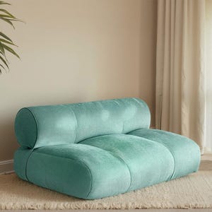 May include: A teal, low-profile sofa with rounded backrests and seat cushions. A woven basket holds a tall green plant. A small wooden side table holds a lamp with a red apple base. The scene is set on a cream rug.