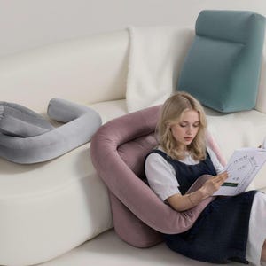 May include: A woman reading a book while seated on a white sofa, supported by a large, U-shaped, dusty rose-colored pillow. A gray, U-shaped pillow and a teal backrest pillow are also on the sofa. The woman is wearing a white shirt and a navy blue dress.