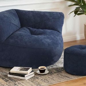 May include: A navy blue sectional sofa with a matching ottoman. The sofa has a rounded corner design and a textured fabric. A small coffee table with a cup of coffee and books is in front of the sofa. A potted plant is in the background.