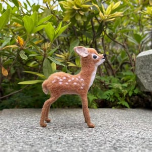Puede incluir: Figura de ciervo de fieltro hecha a mano. El ciervo tiene un cuerpo marrón con manchas blancas, detalles blancos en la cara y las orejas, y patas marrones delgadas. La figura está sobre una superficie gris, con follaje verde en el fondo.