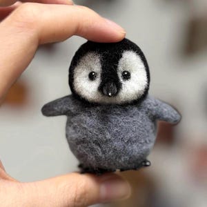 Könnte beinhalten: Eine handgefertigte Pinguinfigur aus Filz. Der Pinguin hat einen schwarzen Kopf und Rücken, mit einem weißen Gesicht und Bauch. Seine Flügel sind klein und grau, und er hat einen kleinen schwarzen Schnabel und Knopfaugen. Der Pinguin ist etwa 7.6 cm groß.