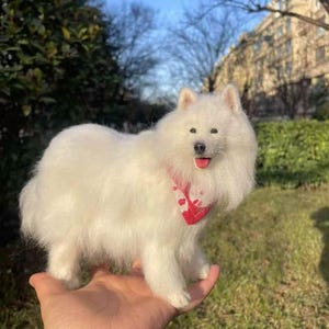 May include: A fluffy, white Samoyed dog figurine with a red patterned bandana around its neck. The dog is standing on a hand, with a green and blue background. The dog's tongue is visible.