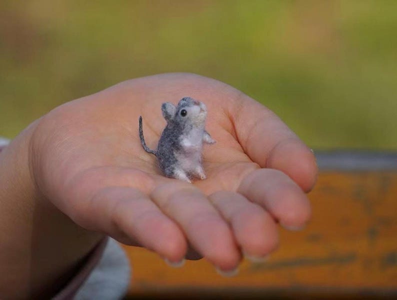 May include: A small, gray and white needle felted mouse is sitting in the palm of a hand. The mouse has a long tail and is looking up.
