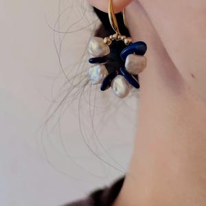 Puede incluir: Pendientes de aro dorados con un racimo de perlas barrocas blancas y detalles de piedras preciosas azul oscuro.
