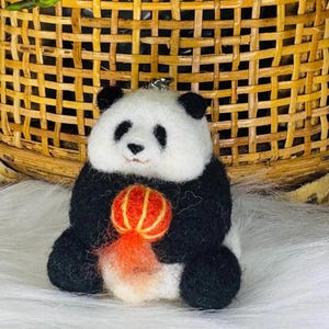 May include: A black and white panda bear holding a red and orange Chinese lantern. The panda is sitting on a white surface.