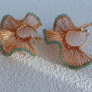 Pode incluir: Um par de brincos feitos à mão com um design único. Os brincos são feitos com arame cor de cobre, moldados em um design floral e adornados com pequenas contas verdes claras. Os brincos têm um aro e um pino dourados.