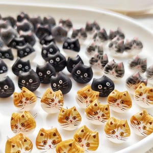 Puede incluir: Una colección de pendientes hechos a mano con forma de gato. Los pendientes están hechos de arcilla polimérica y pintados con varios colores, incluyendo negro, blanco, gris y naranja. Los gatos tienen diferentes expresiones faciales y están dispuestos en círculo sobre un plato blanco.