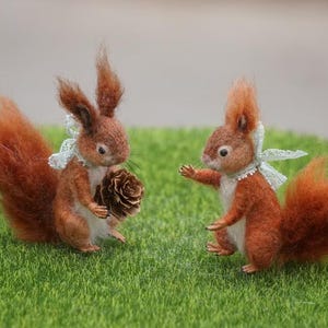 May include: Two brown and white needle felted squirrels with long bushy tails. One squirrel is holding a small brown pine cone. Both squirrels are wearing a white lace collar with a blue ribbon.