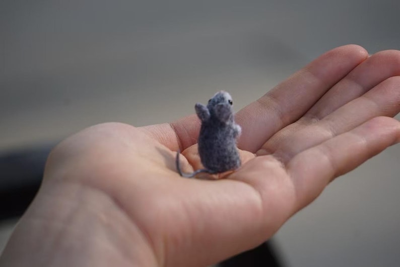 May include: A small gray felt mouse figurine sitting in the palm of a hand.