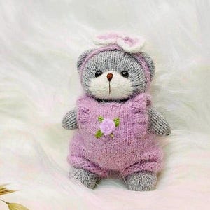 May include: A gray knitted teddy bear wearing a pink knitted outfit with a flower detail. The bear has a white bow on its head.