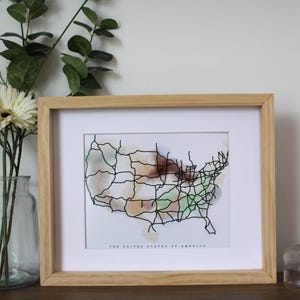 May include: A framed print of a map of the United States with black lines representing roads and highways. The map is overlaid with a watercolor wash in shades of green, brown, and orange. The text "The United States of America" is printed below the map.