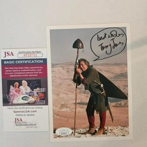May include: A rectangular photograph featuring a person in chainmail armor holding a tall staff with a helmet on top, set against a desert landscape. The image includes a black ink signature and the handwritten text best wishes Terry Jones.