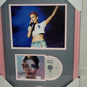 May include: A framed display featuring a photograph of Halsey performing on stage, a smaller portrait of Halsey with a printed signature, and a Manic CD. The frame is pink with a gray mat, and the corners are protected by white cardboard.