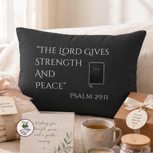 Puede incluir: Cojín decorativo negro con el texto "THE LORD GIVES STRENGTH AND PEACE" y "PSALM 29:11". También presenta una pequeña ilustración de la Biblia. Una tarjeta dice "Wishing you strength, peace, and a gentle recovery."
