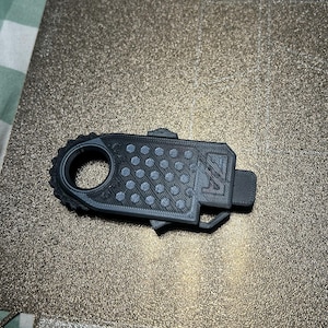 Puede incluir: Un objeto gris oscuro impreso en 3D con un extremo circular en forma de engranaje y un cuerpo rectangular con una serie de pequeñas hendiduras circulares de color claro. El objeto tiene una pequeña extensión rectangular.