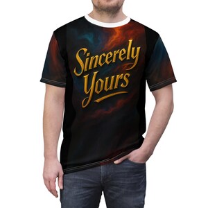 以下が含まれることがあります： 「Sincerely Yours」の金色の文字が入った黒いTシャツ。白いクルーネックと半袖です。シャツの背景には、オレンジと青の色合いの暗く曇ったデザインが施されています。