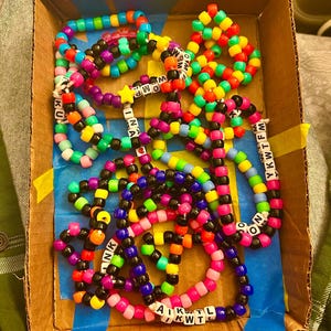 Può includere: Una collezione di braccialetti colorati con perline e lettere che compongono diverse parole, tra cui "WOM", "YKWTFM", "INA", "KU", "NK", "AKTL" e "KWT".