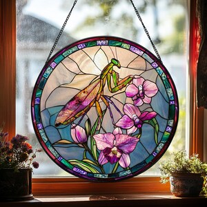 May include: Round stained glass window art featuring a praying mantis and orchids. The mantis is depicted in shades of green, yellow, and purple, with the orchids in pink and purple. The window is framed with a colorful border and hangs from a chain.