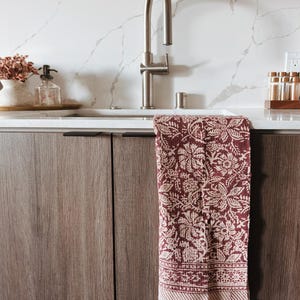 May include: A burgundy and cream floral patterned kitchen towel hangs from a stainless steel faucet. The towel has a fringed edge and is draped over a white sink. The background features a marble-look backsplash and wooden cabinets.