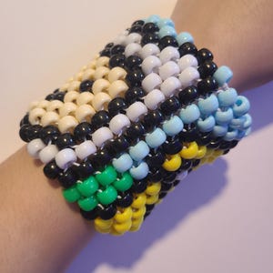 May include: A colourful beaded bracelet featuring a pattern of black, white, yellow, green, and blue beads. The bracelet is made of small, round beads strung together to form a wide cuff. The bracelet is approximately 7.6 cm wide.